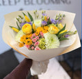 Sunny Days Bouquet featuring yellow ranunculus, tulips, sunflowers, pink spray roses, white chrysanthemums, lavender lisianthus, and hydrangea wrapped in cream paper