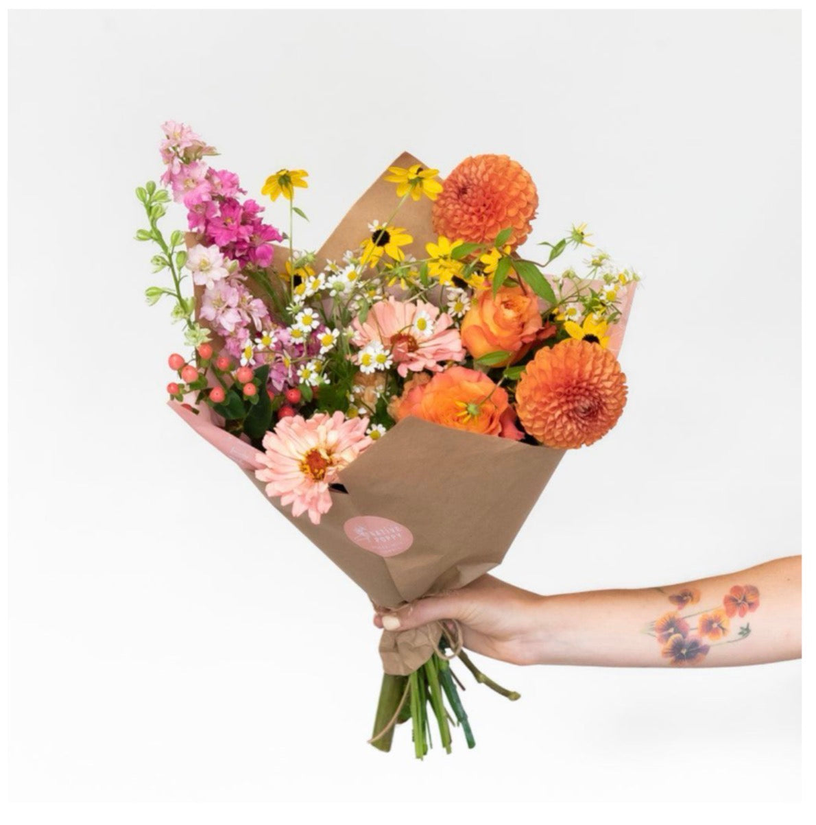 Golden Hour Bouquet featuring orange dahlias, ranunculus, pink gerbera daisies, yellow rudbeckia, chamomile, stock flowers and hypericum berries in kraft paper wrap